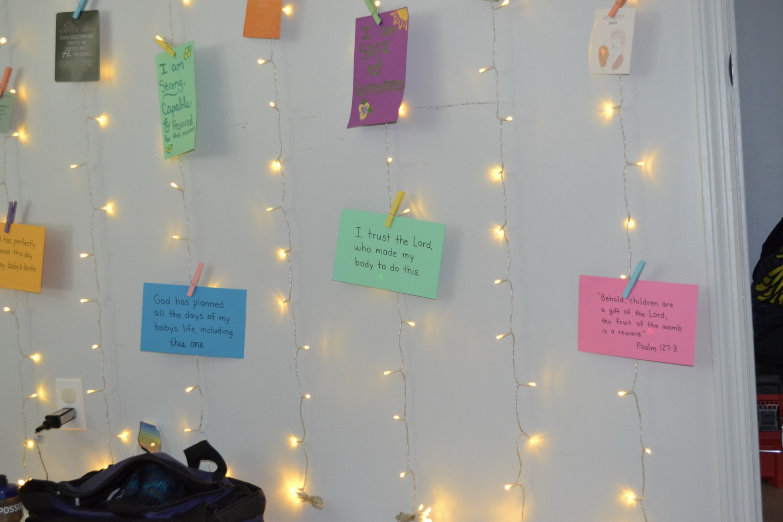 A wall of handwritten birth affirmation cards pegged to strings of warm fairy lights, one of them reading &lsquo;I trust the Lord, who made my body to do this&rsquo;.