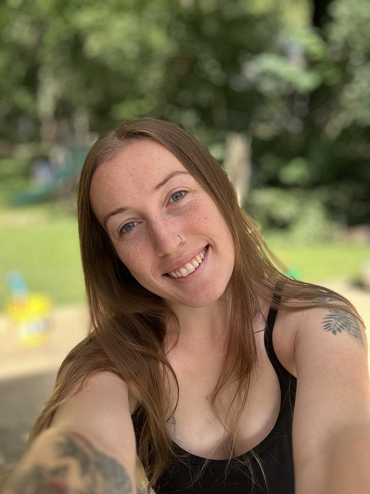 Megan Quintanilla smiling outdoors, with long auburn hair and a black tank top, trees and grass blurred behind her.