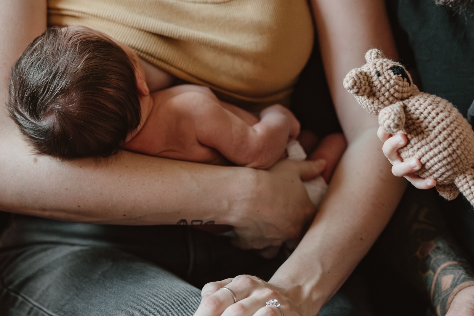 A newborn rests skin-to-skin on their mother&rsquo;s chest while a small crocheted bear is held out beside them.