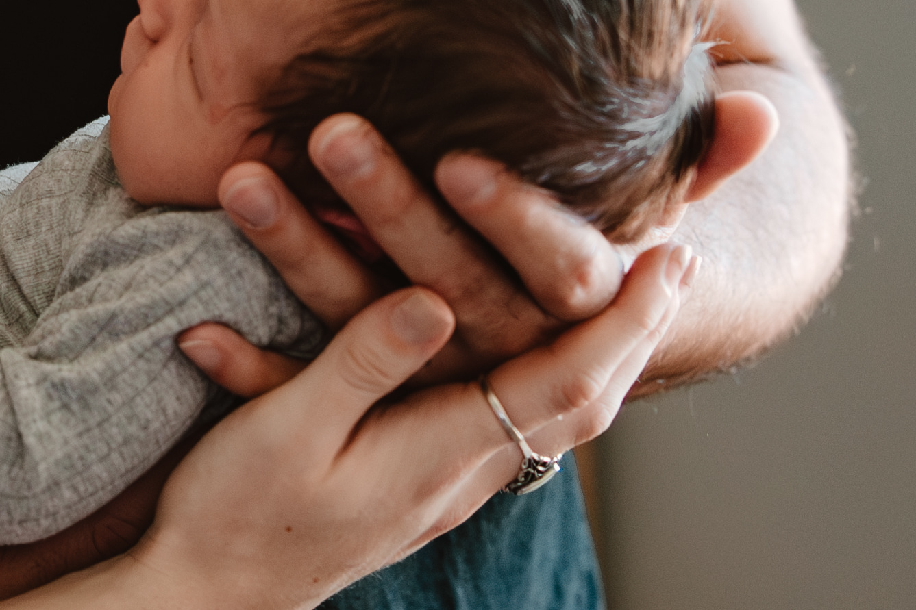 A newborn resting in an adult&rsquo;s hands, one hand cupped behind the head and another supporting the body.
